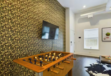 From this angle, the living room shines with a bold camouflage accent wall, a vibrant foosball table, & playful framed prints. The hypebeast-themed rug adds character, complementing the modern decor & creating a dynamic yet cozy entertainment space.