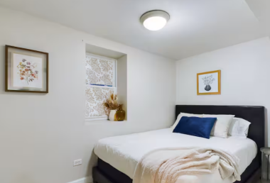 Enjoy restful nights in this bright, inviting bedroom featuring a luxurious queen bed, soft pillows, and a stylish throw. Framed art and a charming window with patterned curtains add a personal touch to the cozy ambiance.