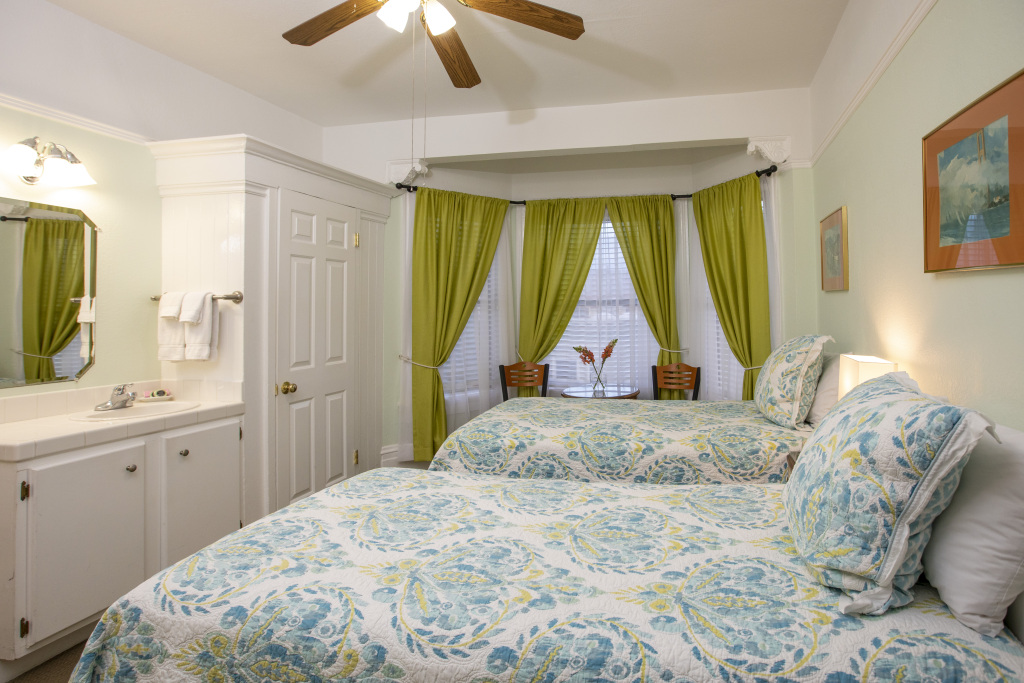 A hotel room with two beds, a ceiling fan, green curtains, a white cabinet, and a mirror.