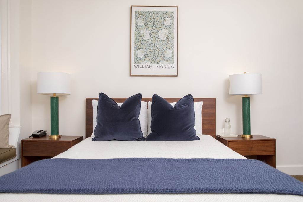 A neatly made bed with a blue blanket, two nightstands with lamps, and a piece of artwork above the bed.
