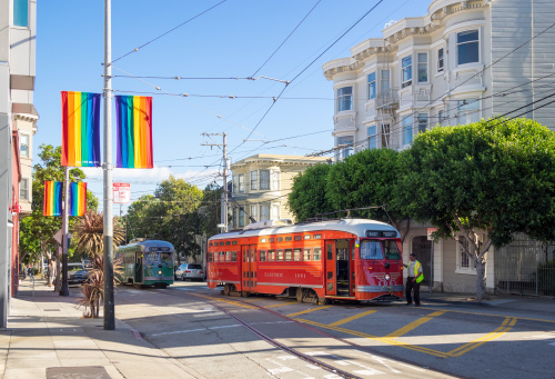 A street scene with a red trolley, rainbow flags, and buildings.