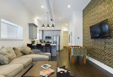 This vibrant living room features a plush sectional couch for ultimate relaxation, a sleek 55-inch TV for entertainment, & a trendy foosball table for fun. The eye-catching hypebeast-inspired accent wall adds personality to this modern space.