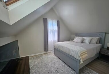 Bedroom 2 offers a cozy queen bed with soft linens and plush pillows. Natural light streams through the skylight and arched window with curtains. A 40