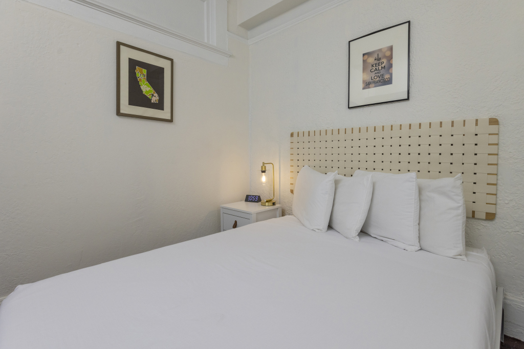 A neatly made bed with white linens and pillows in a room with white walls and framed pictures.