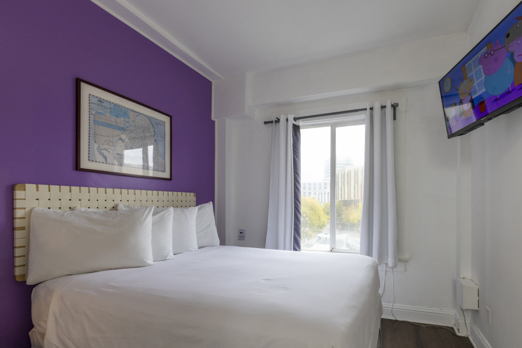 A hotel room with a large bed, window, and TV on the wall.