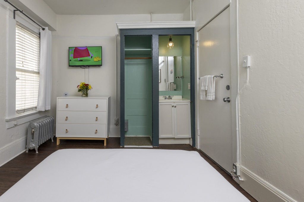 A hotel room with a white bed, dresser, TV, window, and bathroom