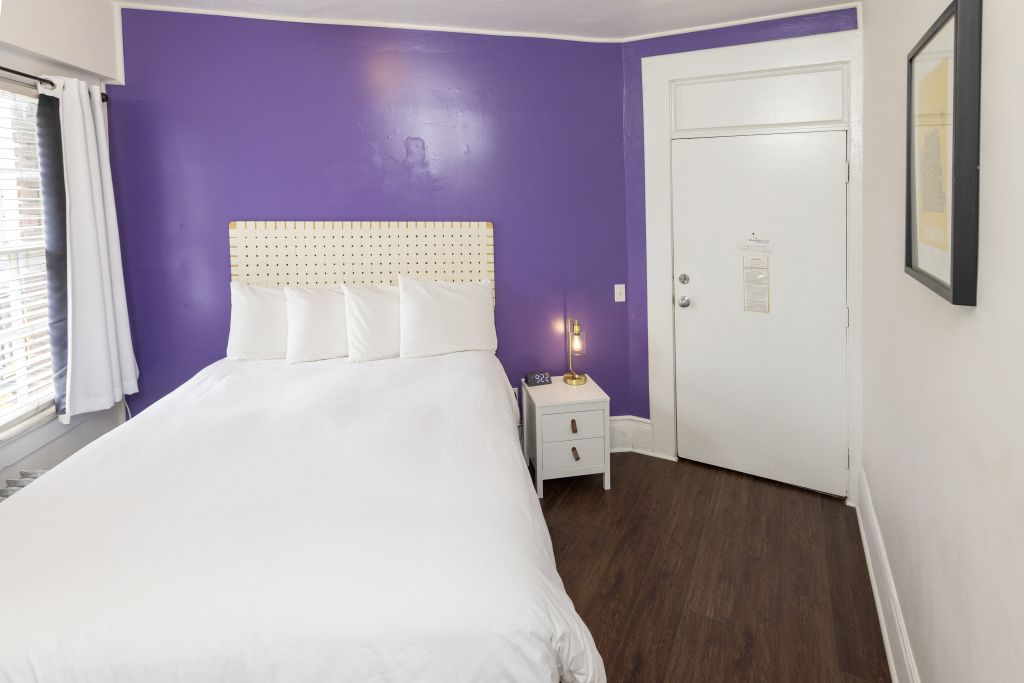 A hotel room with a purple accent wall, a white bed with a beige headboard, a nightstand with a lamp, and a white door.
