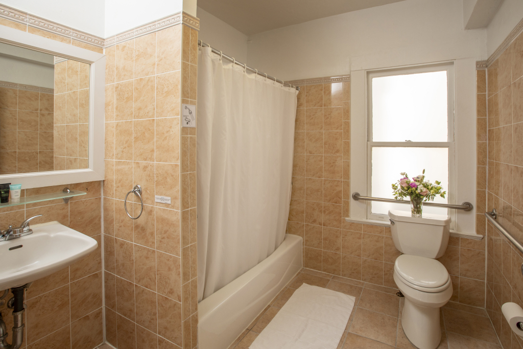 A clean bathroom with tan tiles, a white sink, toilet, and shower with a curtain, and a window with flowers on the toilet