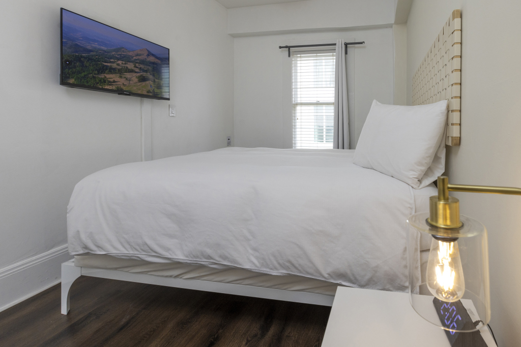 A neatly made bed with white linens and a flat-screen TV mounted on the wall in a simple bedroom with a window.