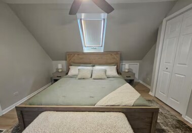 From the foot of the bed, admire the elegant wooden headboard framed by a skylight offering natural light, with blinds for a cozy ambiance. The cushioned bench adds charm, while matching side tables with chic lamps create a balanced, tranquil vibe.