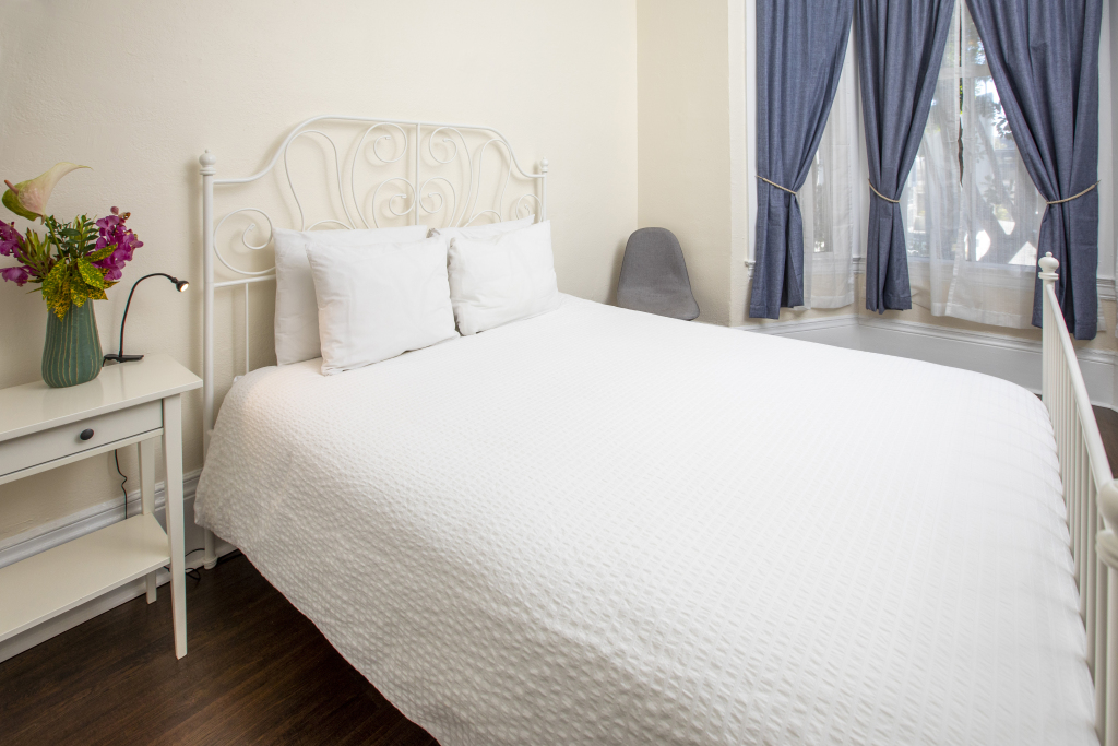 A neatly made bed with white linens and pillows in a room with a window, curtains, and a nightstand with flowers.