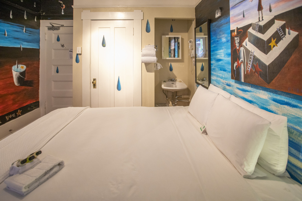 A wide-angle view of a hotel room with a large bed, murals on the walls, and a sink area.