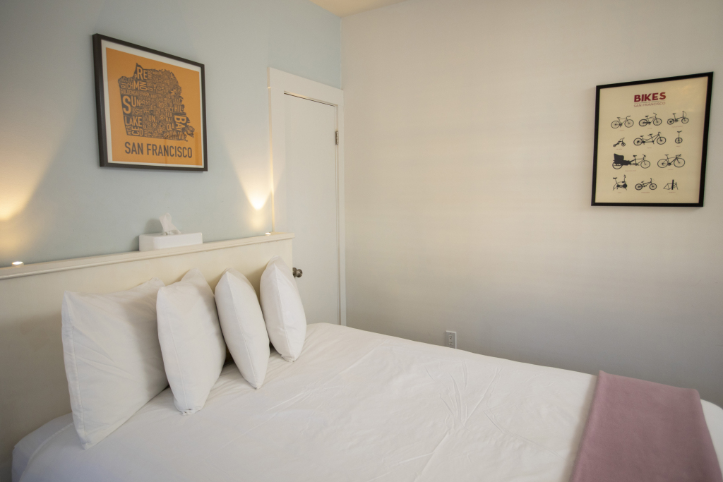 A neatly made bed with white linens and pillows in a room with light blue walls, two framed posters, and a tissue box on a shelf.