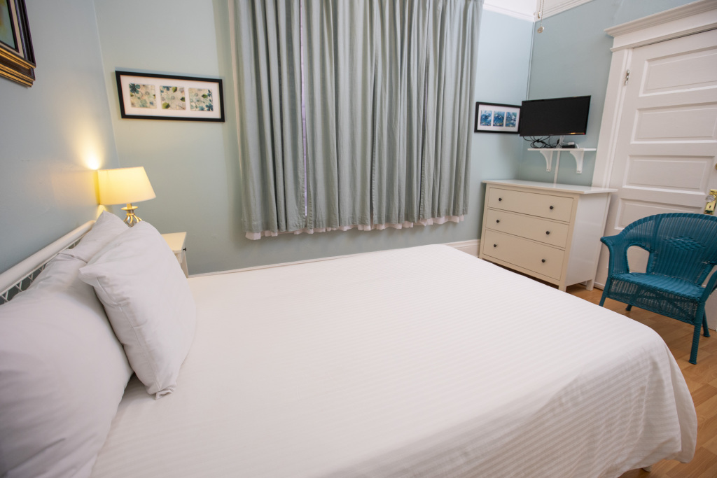 A neatly made bed with white linens and pillows, a blue wicker chair, a white dresser with a TV on top, and a window with curtains in a room with light blue walls.