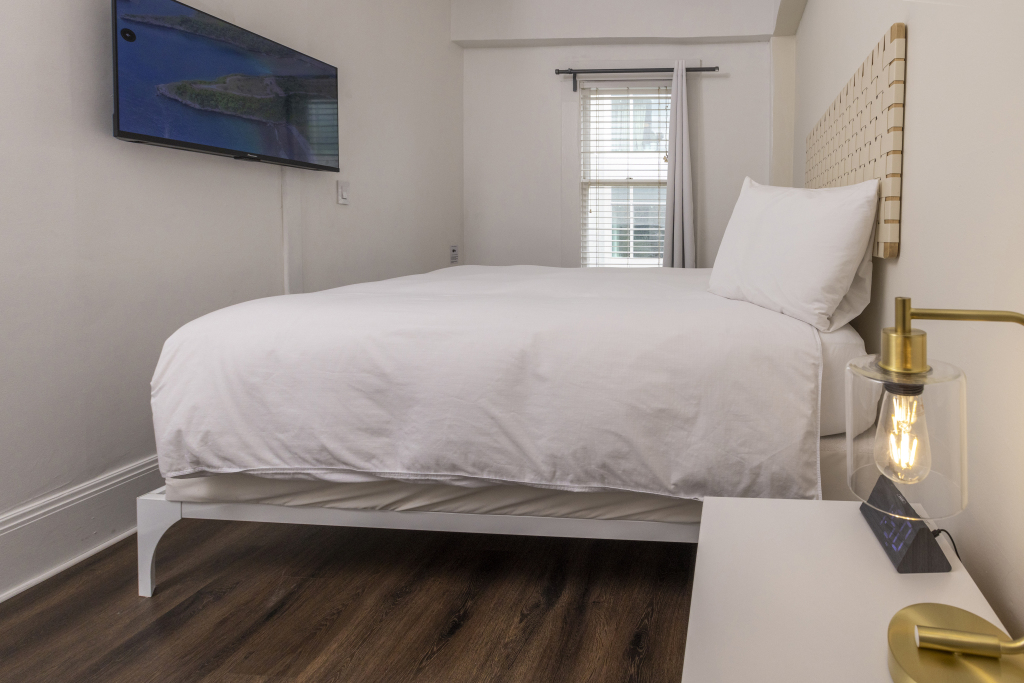 A neatly made bed with white linens and a flat-screen TV mounted on the wall in a simple room with a window.