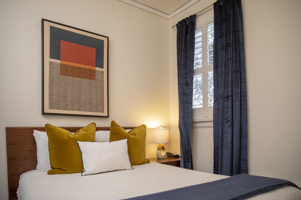 A hotel room with a bed, window, artwork, and nightstand.