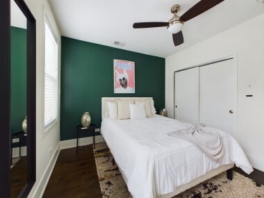 Bedroom 1 features a plush white bed with a sleek headboard, a striking green accent wall, and vibrant wall art. A large window allows natural light, while a full-length mirror and minimalist furniture create a serene and stylish atmosphere.