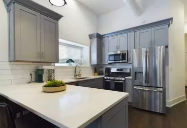 This full kitchen is a chef's dream, with stainless steel appliances, including a refrigerator, oven, & microwave. The wide quartz countertop doubles as a prep & dining area, complemented by sleek grey cabinetry, a deep sink, & a bright window view.