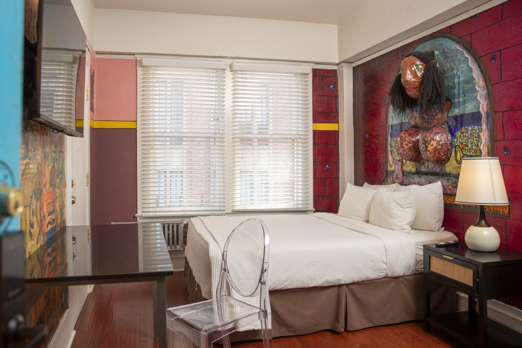 A hotel room with a bed, desk, and chair in front of a window with colorful artwork on the walls.