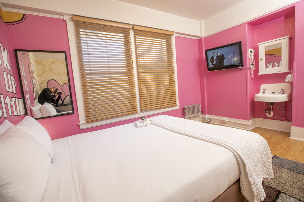 A wide view of a hotel room with pink walls, a white bed, a window with blinds, a flat screen TV, and a small sink with a mirror.