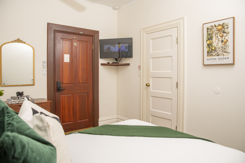 A hotel room with a bed, TV, door, mirror, and artwork.