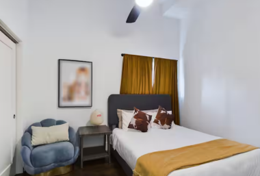 Cozy bedroom featuring a queen bed with stylish pillows and a mustard throw for a pop of color. The room is accented with warm gold curtains and modern decor. A ceiling fan provides comfort, while the closet offers ample storage for your belongings.