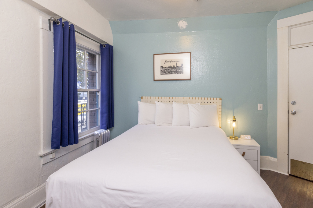 A neatly made bed with white linens and pillows against a light blue wall with a window and a framed picture above the bed.