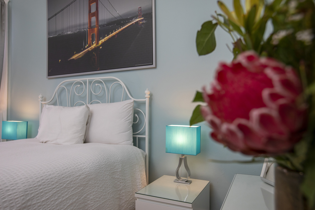 A neatly made bed with white linens and a white metal frame, with a blue lamp and a picture of the Golden Gate Bridge on the wall.