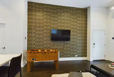 The living room boasts a striking feature wall with bold, modern design, perfectly framing the sleek wall-mounted TV. A foosball table invites fun, while the rich wood flooring and minimalist table setups complete this dynamic and inviting space.