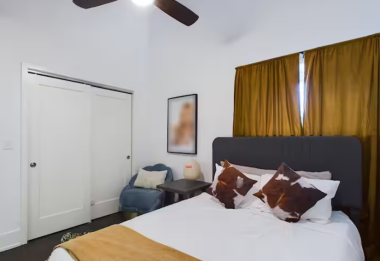 This bedroom features a plush bed with soft linens, a sleek headboard, golden curtains, and stylish framed decor. A cozy chair, modern side table, lamp, ceiling fan, and sliding doors complete the warm, inviting space.