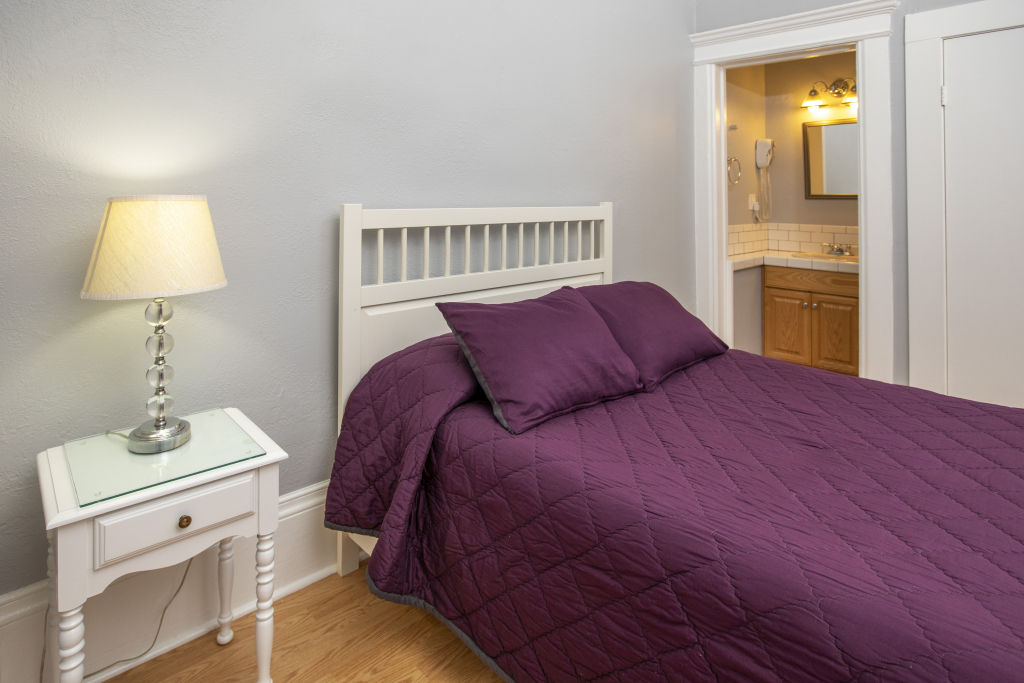 A bedroom with a purple bedspread, a white nightstand with a lamp, and a doorway to a bathroom.