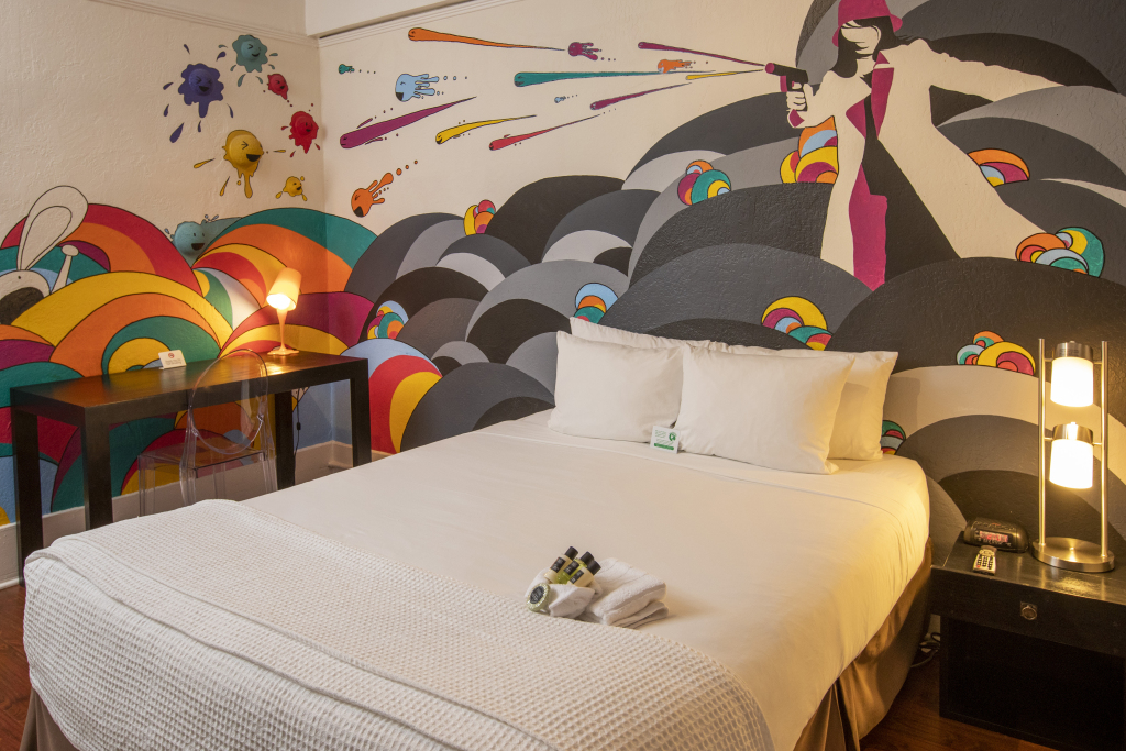 A hotel room with a large bed, desk, and colorful mural on the wall.