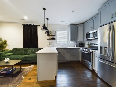 This full kitchen features sleek grey cabinetry, stainless steel appliances, and a bright quartz countertop. The cozy corner layout includes a gas range, microwave, and a double-door refrigerator. A large window adds natural light to the space.