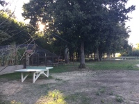 Links befindet sich die XXL-Voliere des Parks mit kleinen Bauernhoftieren.  Ein Klettergerüst und eine Tischtennisplatte. Hinten: Der durch einen Zaun gesicherte Angelteich mit Fußgängertor.
