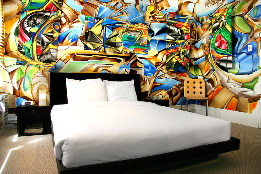 A hotel room with a large abstract art mural on the wall, a bed, nightstand, and chair.