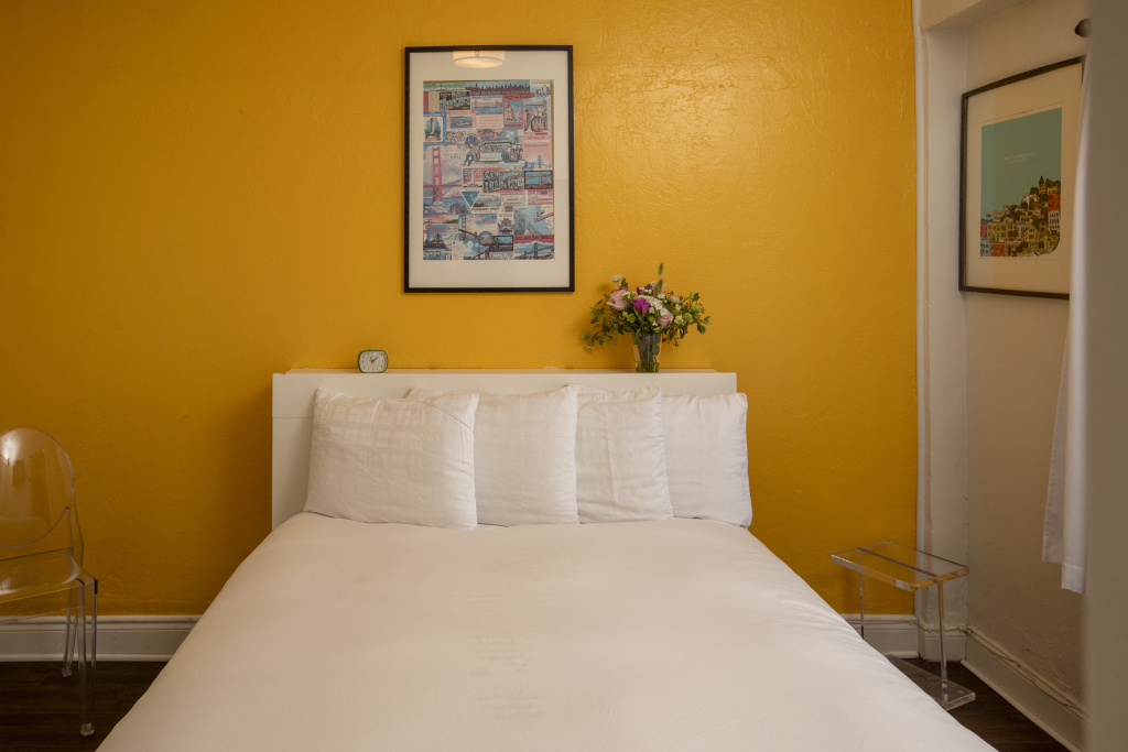 A neatly made bed with white linens and pillows against a yellow wall with a framed picture and a vase of flowers on the headboard.