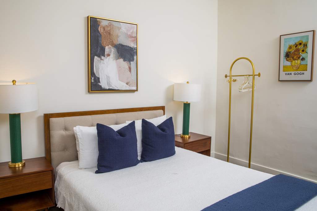 A neatly made bed with pillows and a blue blanket in a room with white walls, two nightstands with lamps, and framed artwork.
