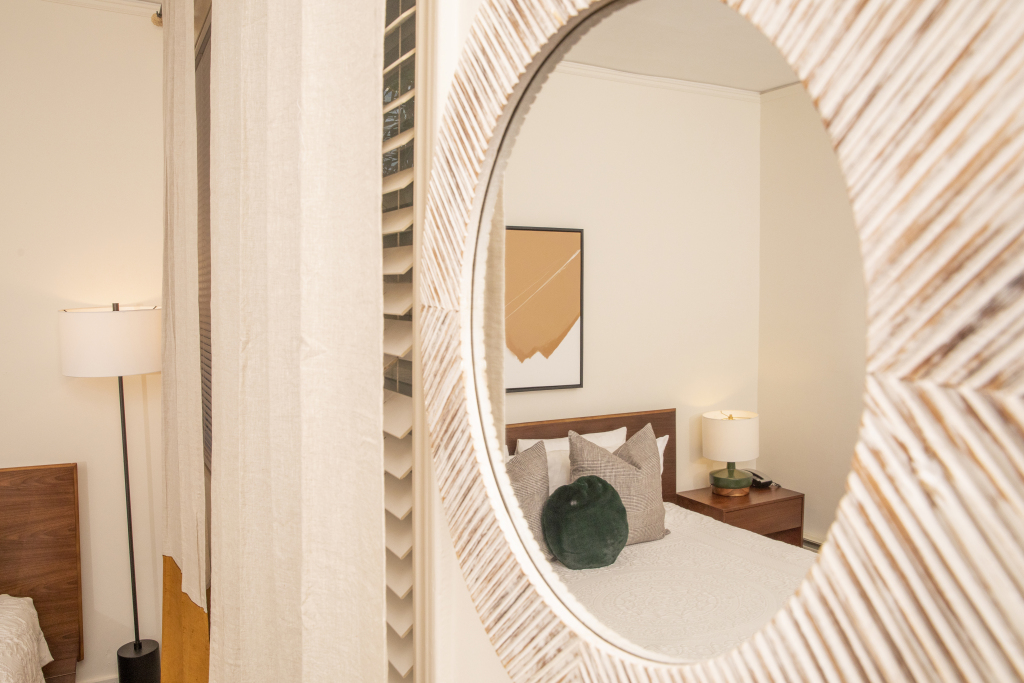 A hotel room with a bed, nightstand, lamp, and artwork reflected in a round mirror.