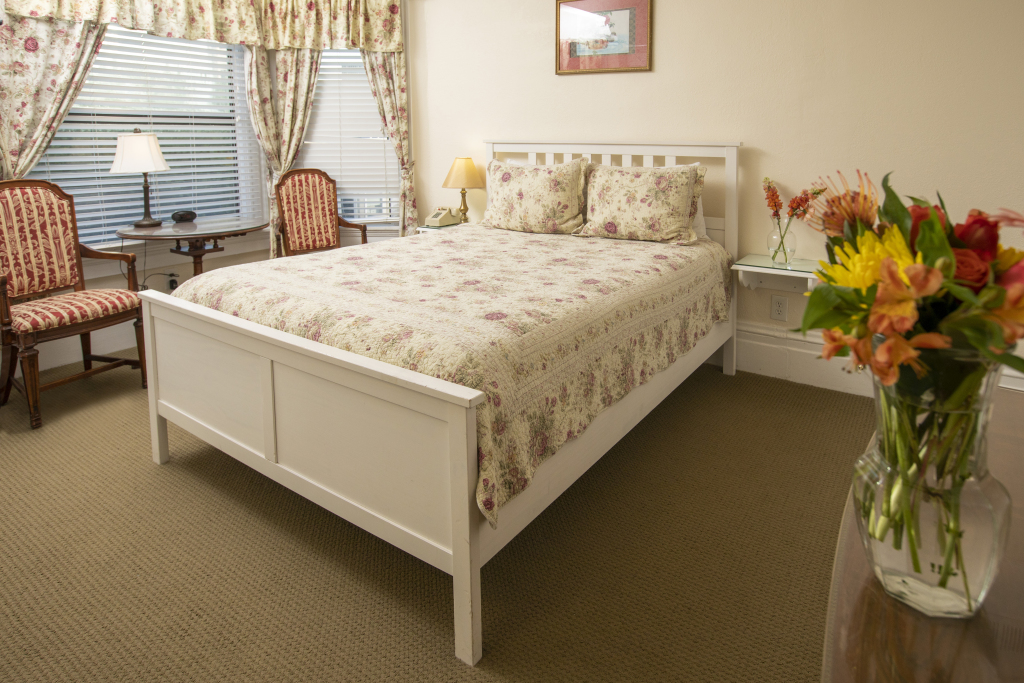 A neatly made bed with floral bedding in a room with a window, two chairs, and a lamp.