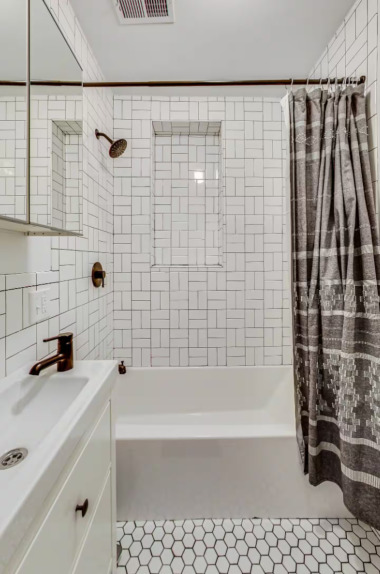 Relax in this elegant bathroom featuring a sleek tub with a stylish tiled surround, a modern sink with bronze fixtures, and ample storage in the mirrored cabinet. The soft gray shower curtain adds a touch of sophistication to this calming space.
