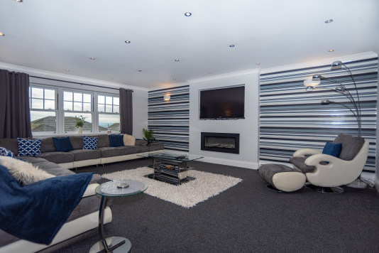 Expansive living room with generous seating for groups, a 4K smart TV, and a feature fireplace for cosy winter evenings, all framed by beautiful sea views.