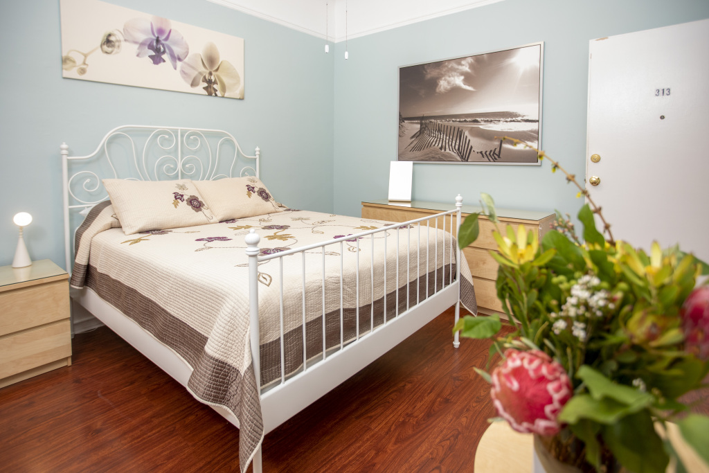 A tidy hotel room with a white bed, nightstand, and lamp, featuring light blue walls and a large piece of beach-themed artwork.