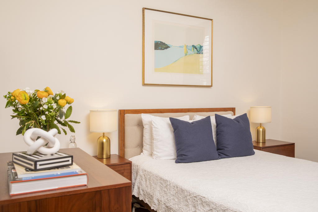 A neatly made bed with white and blue pillows, flanked by nightstands with lamps and a vase of yellow flowers on one side.