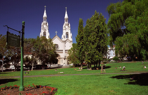 a white church with two towers stands behind a green park with people on the grass