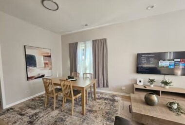 Dine in comfort at this chic wooden table with seating for four, set on a stylish area rug. Enjoy natural light from the window with soft curtains, bold wall art, and the convenience of a TV visible from the adjacent living area for relaxed meals.