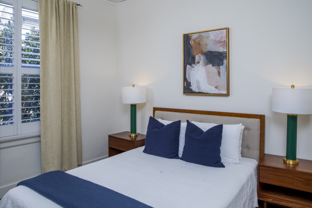 A neatly made bed with blue and white pillows and a blue blanket at the foot, flanked by two wooden nightstands with green lamps, in a room with white walls and a window with beige curtains.