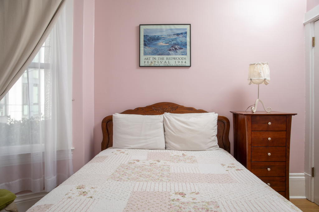 A neatly made bed with white linens and pillows against a pink wall, with a wooden dresser and lamp beside it.
