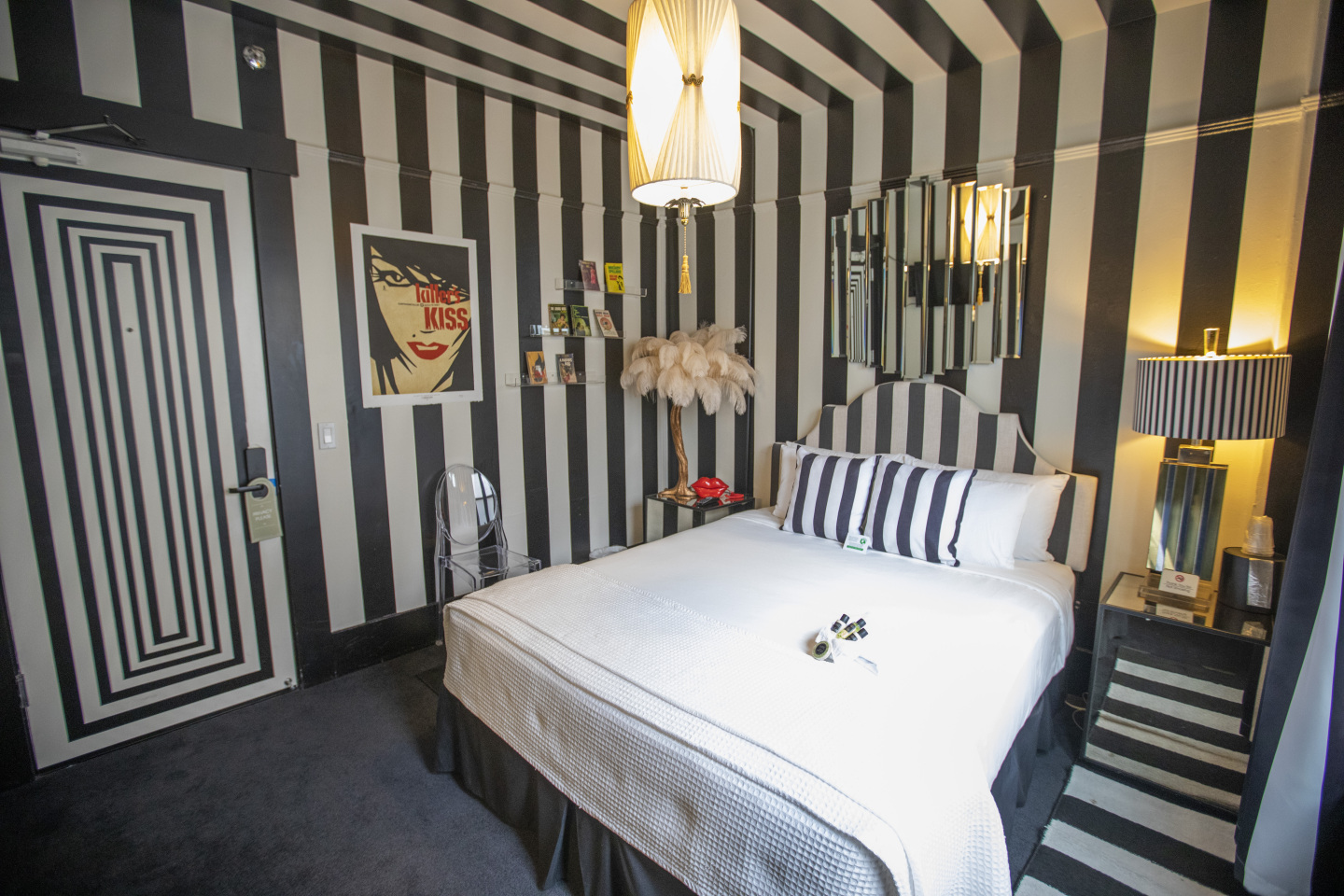 A wide view of a room with a bed, lamp, mirror, and poster on striped walls.