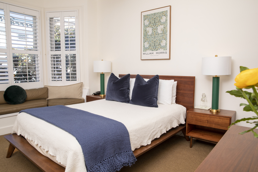 A neatly made bed with a blue throw blanket and several pillows in a room with white walls and a window with shutters.
