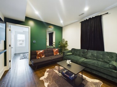 This living room features a striking green accent wall, a sleek mirror with elegant lighting, & a cozy sectional sofa. The cowhide rug adds a touch of rustic charm, while modern bar stools at the counter create a seamless connection to dining area.